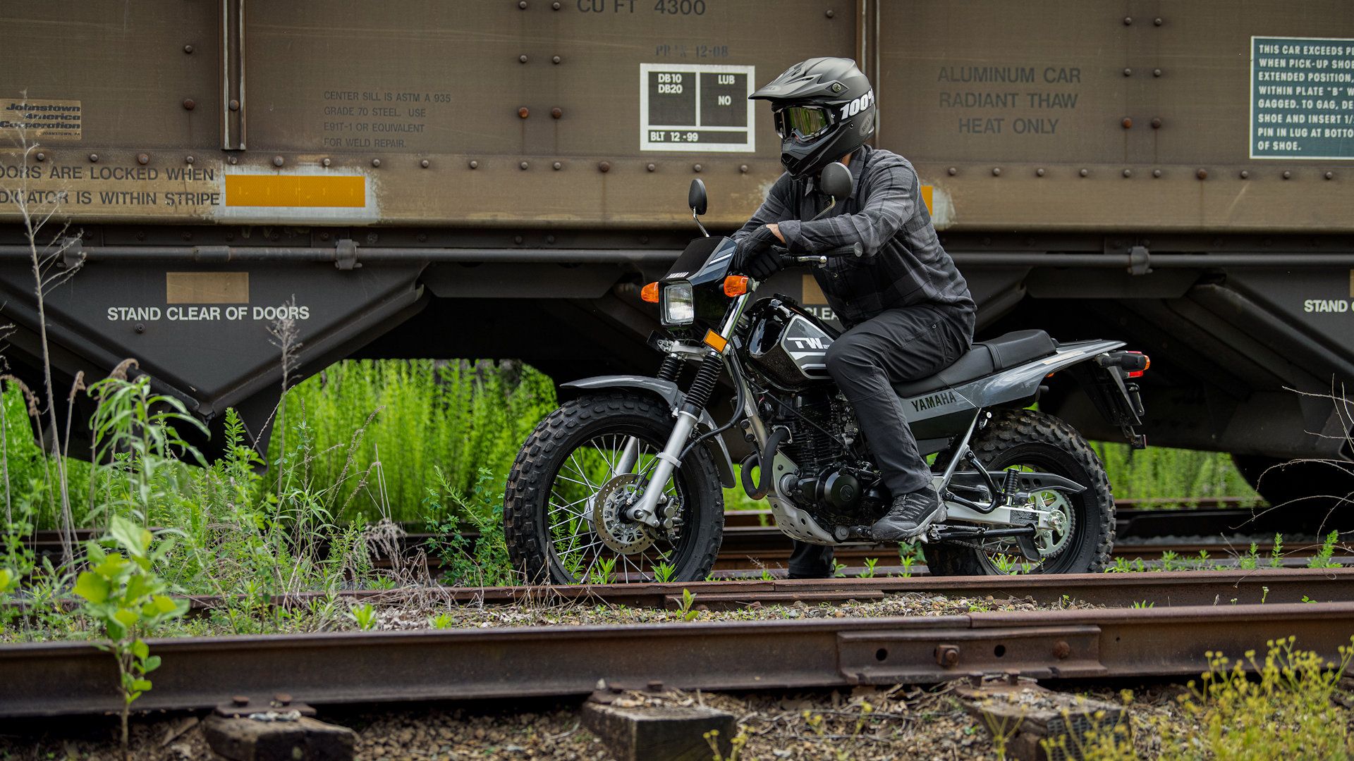 A rider astride a gray 2023 Yamaha TW200 by railroad tracks