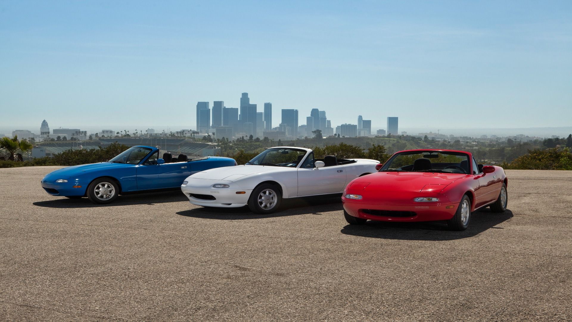 Três Mazda Mx-5 miata, um vermelho, um branco e um azul estacionados.