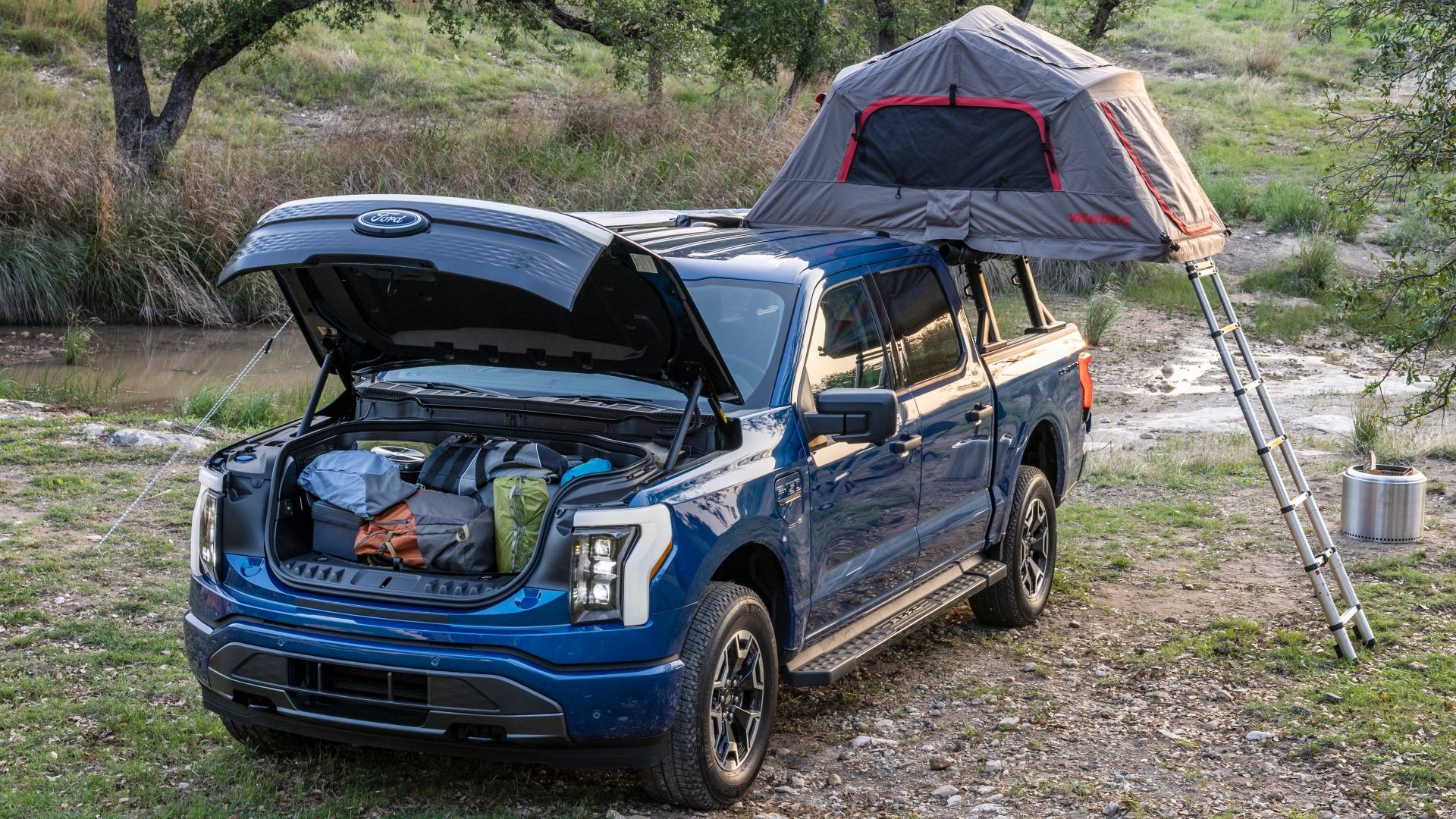 A blue-colored Ford F-150 Lightning is shown with its front trunk fully loaded and a tent attached to its bed at a campsite