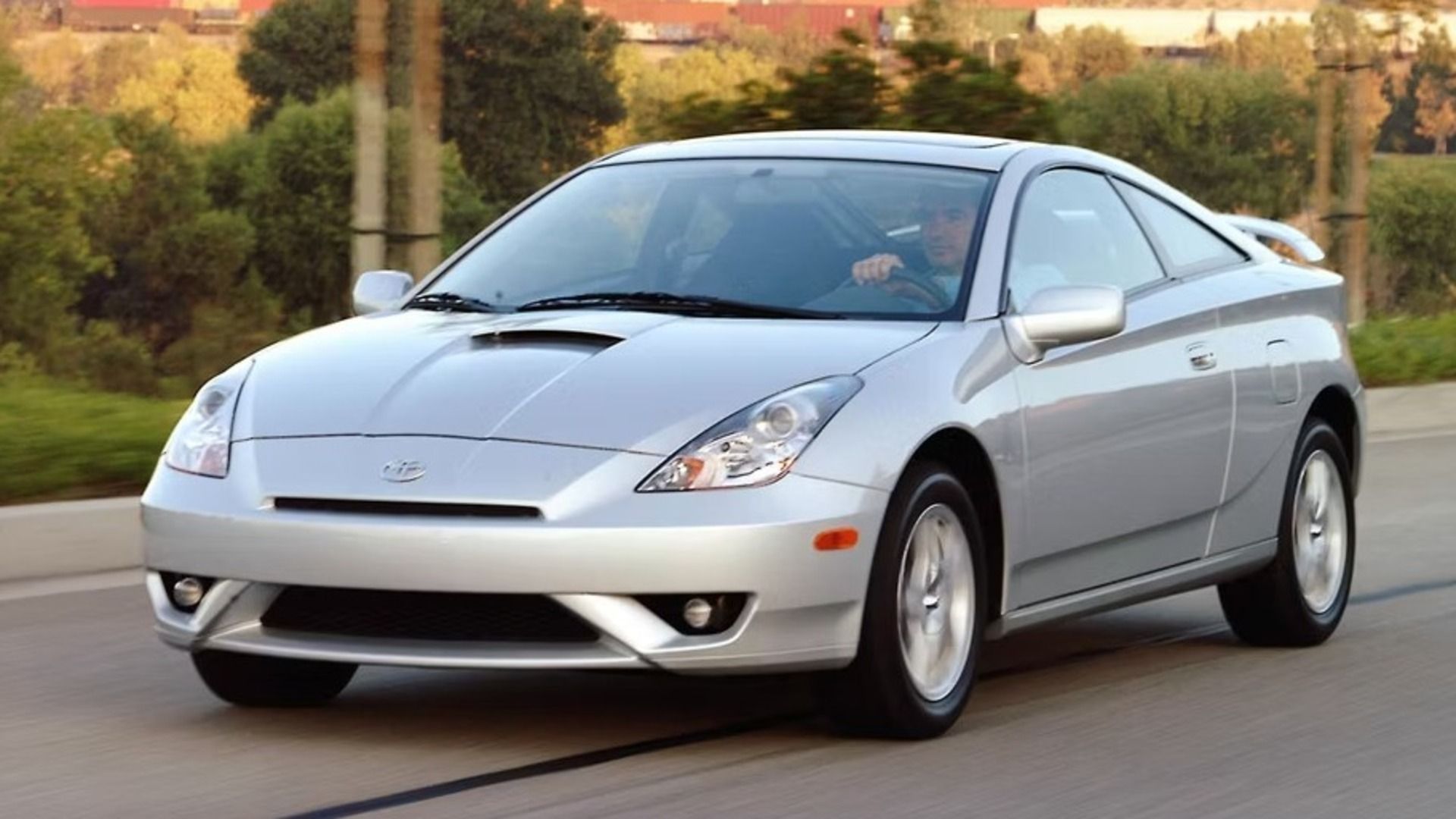 A Silver Toyota Celica T230 on the road driving past trees and bushes.