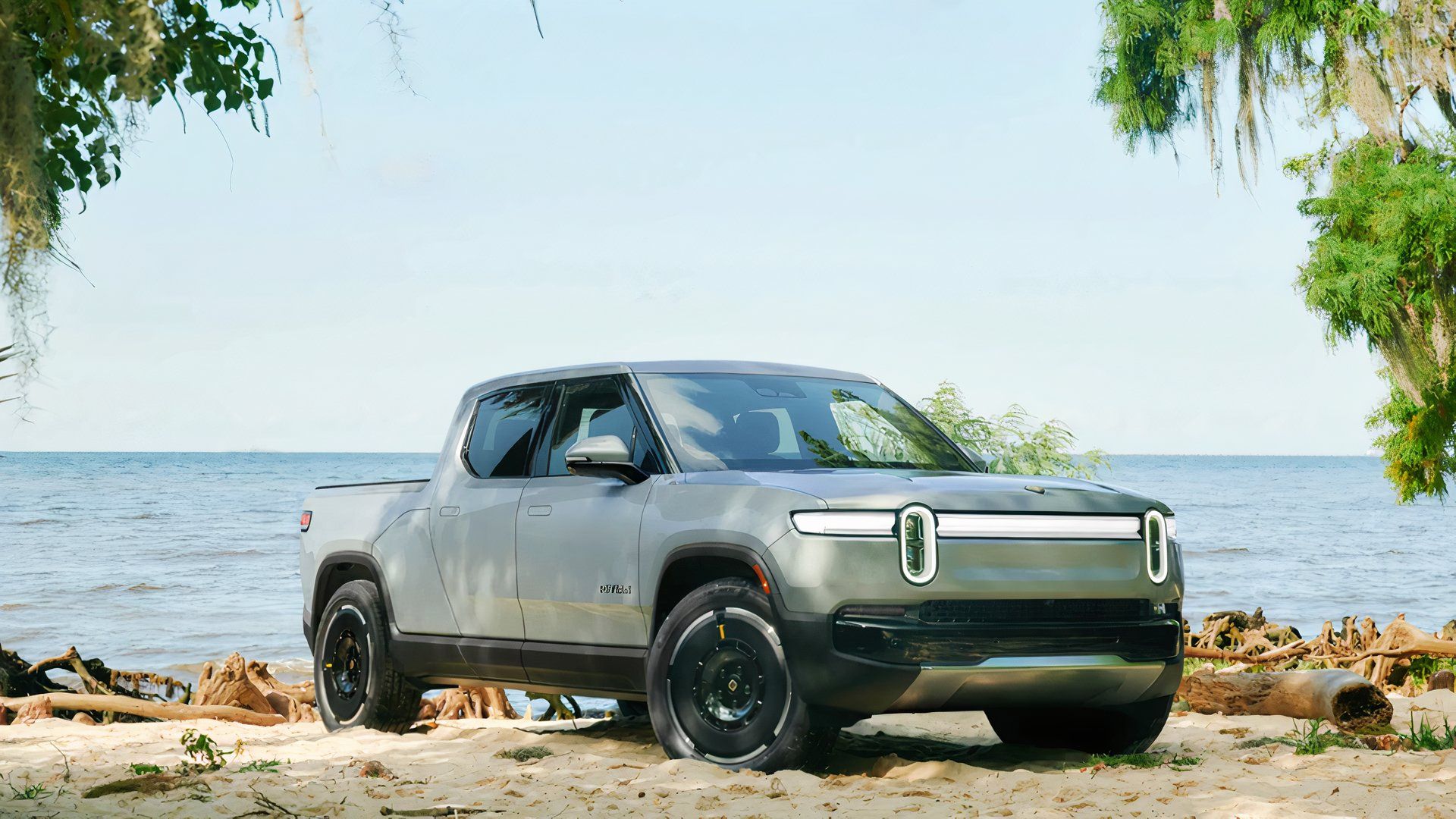 An LA Silver Rivian R1T electric pickup truck is parked on a sandy beach. 