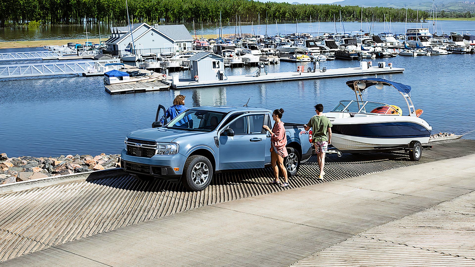 A blue 2024 Ford Maverick Hybrid is towing a boat near water.
