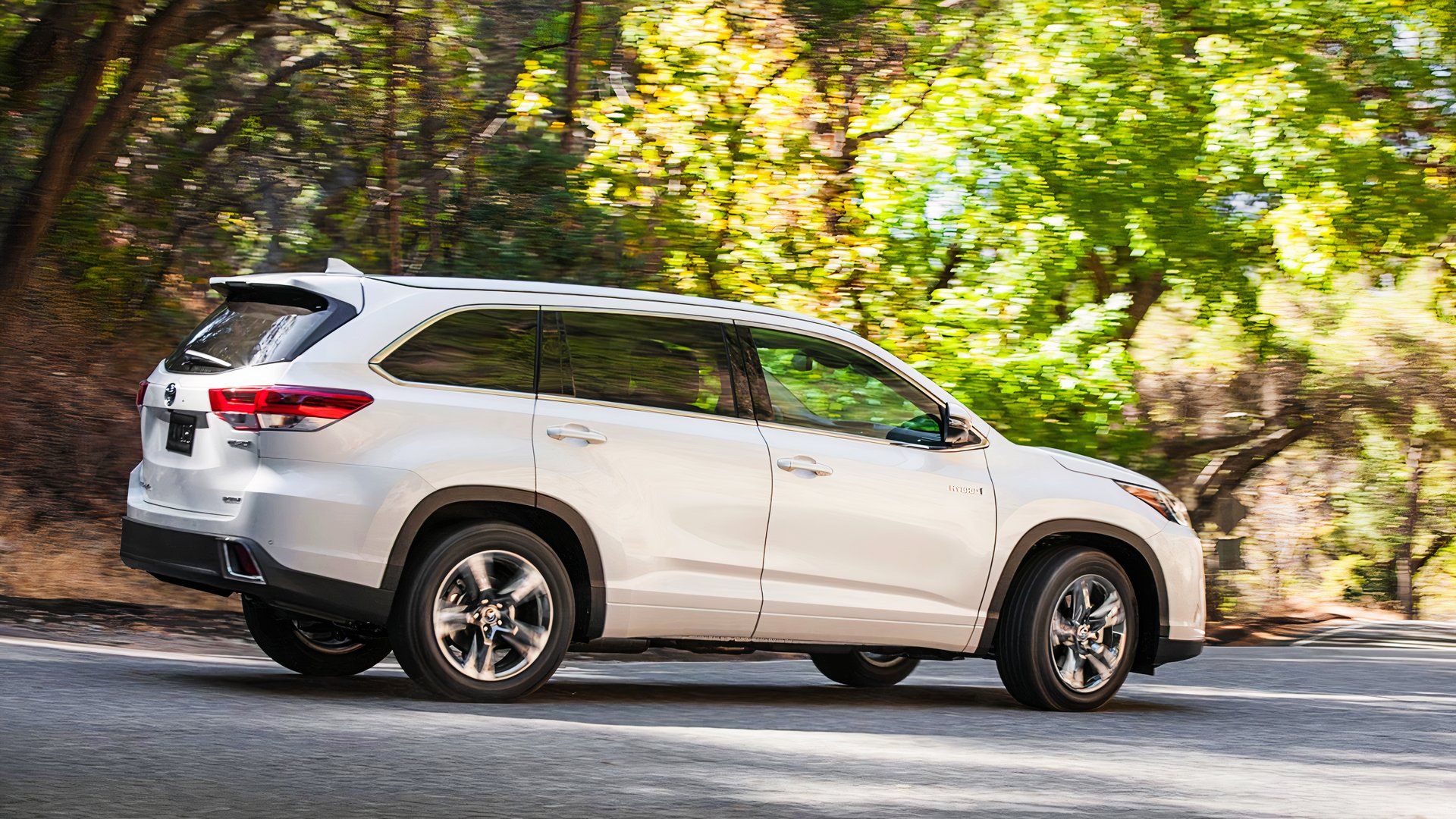 A white 2019 Toyota Highlander Hybrid midsize hybrid SUV is driving. 