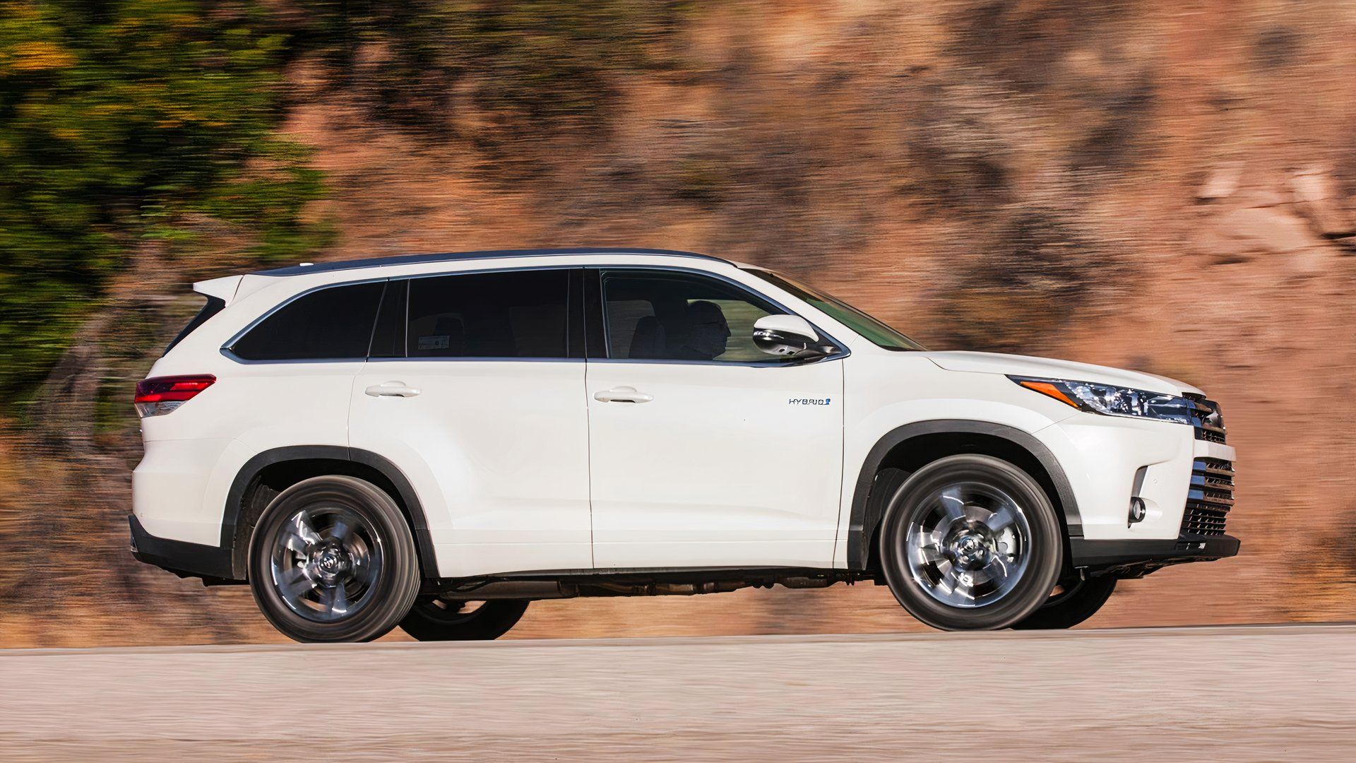 A white 2019 Toyota Highlander Hybrid midsize hybrid SUV is driving. 