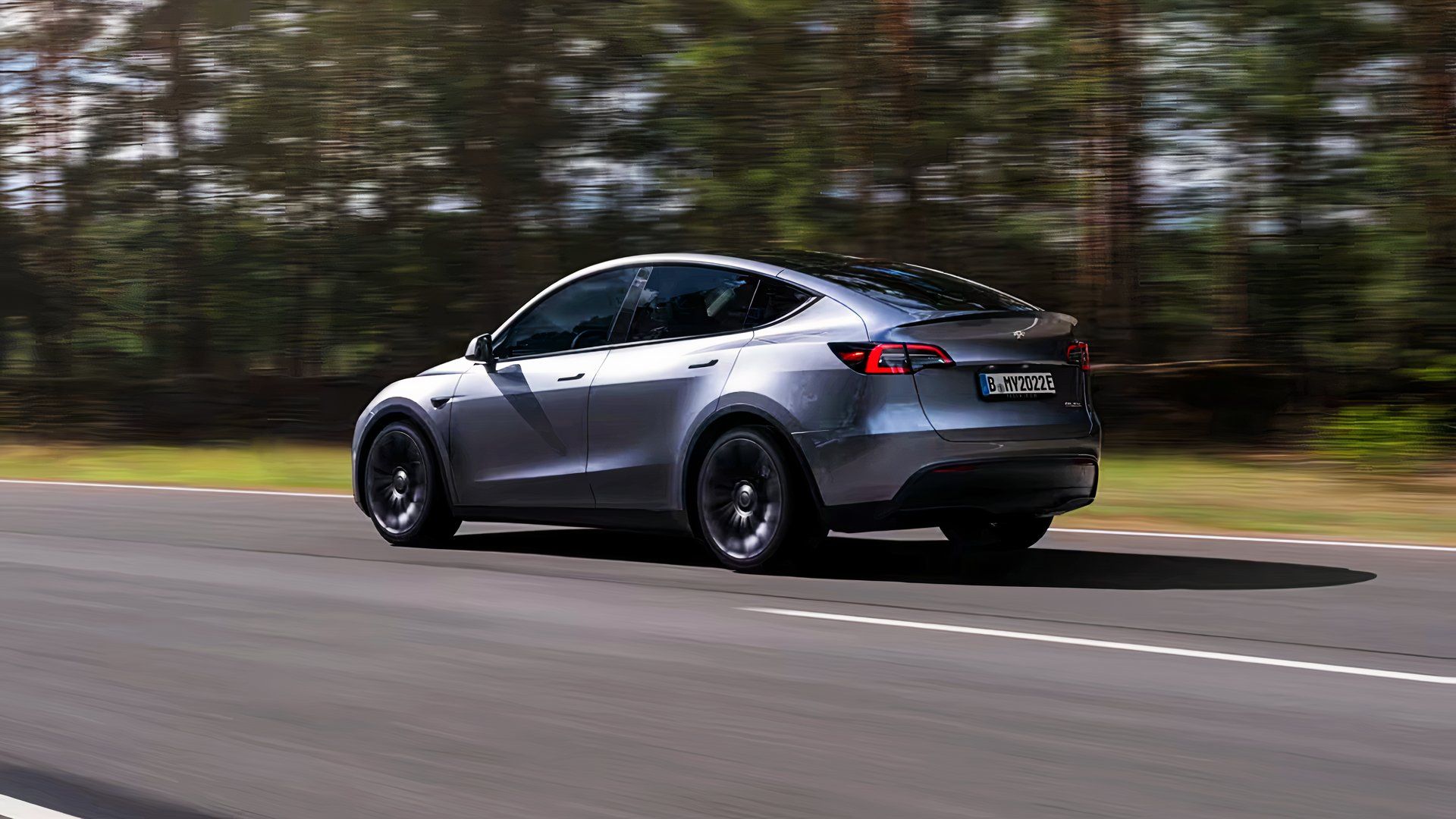 A gray 2024 Tesla Model Y small electric SUV is driving on the road. 