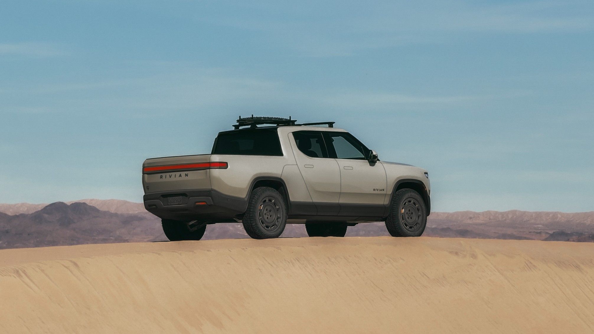 Exterior front three-quarters static image of a gray 2025 Rivian R1T California Dune Edition​​​​​​​​​​​​​​ parked on a sand dune