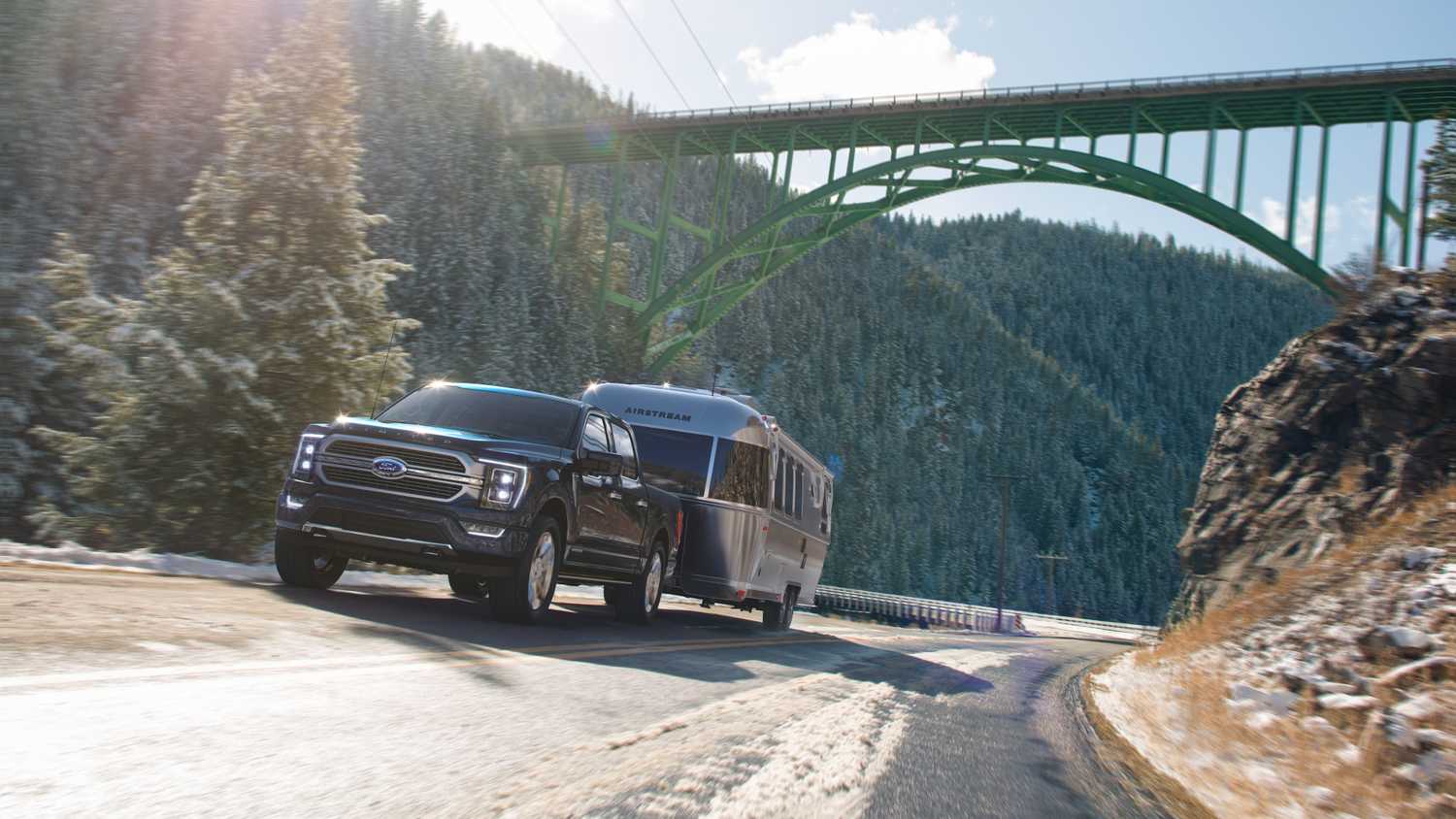 A hybrid pickup truck towing an Airstream trailer through a snowy mountain pass under a green bridge.