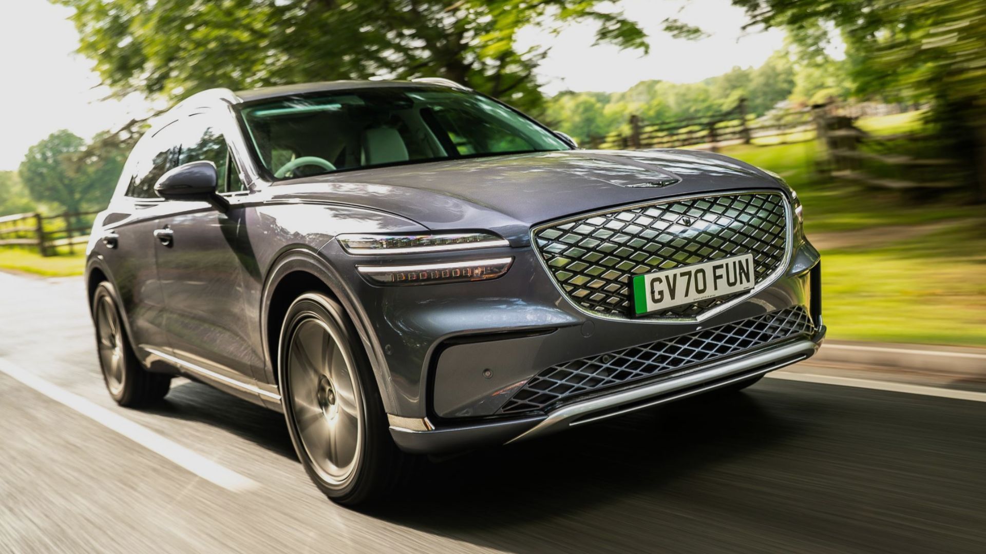 Dynamic front 3/4 shot of a gray 2026 Genesis Electrified GV70 driving on a country road lined by trees.