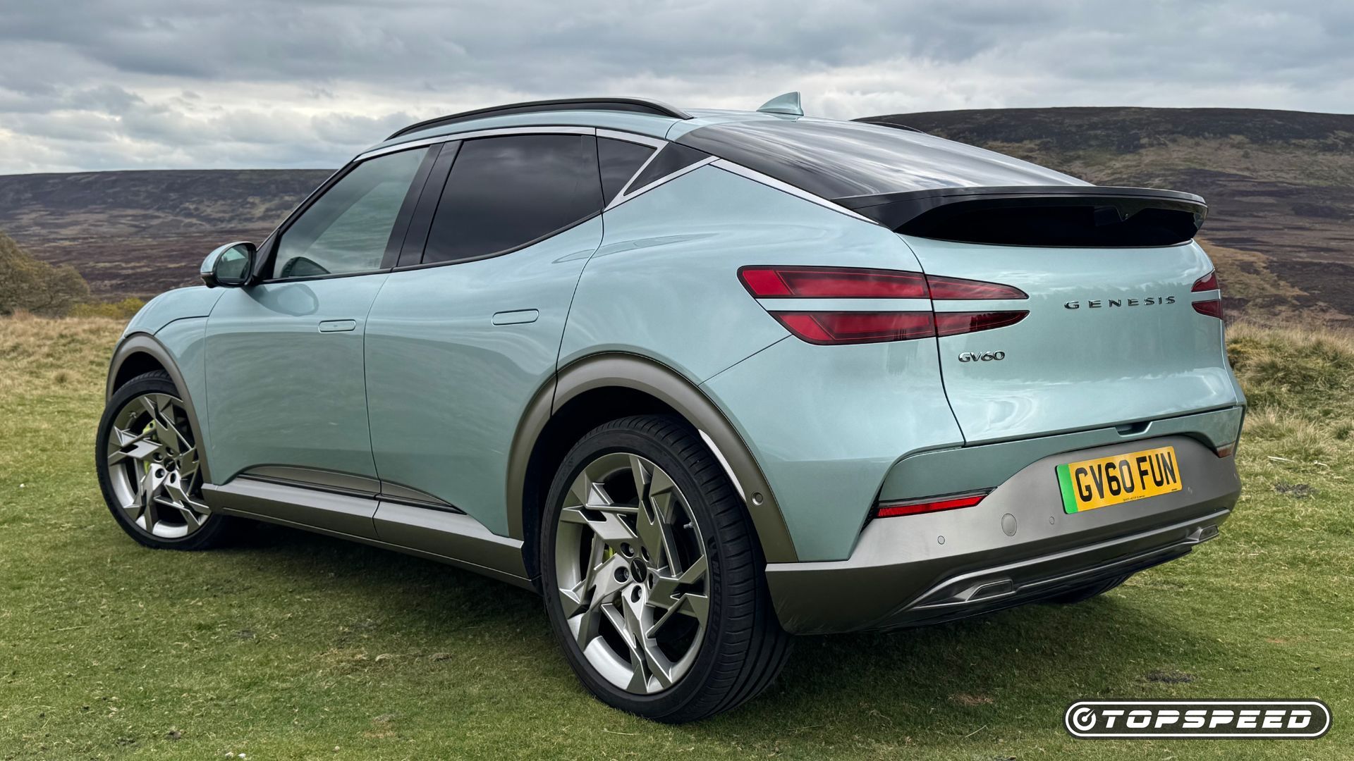 Static rear 3/4 shot of a mint green 2024 Genesis GV60 Sport Plus AWD parked on grass with hills in the background.