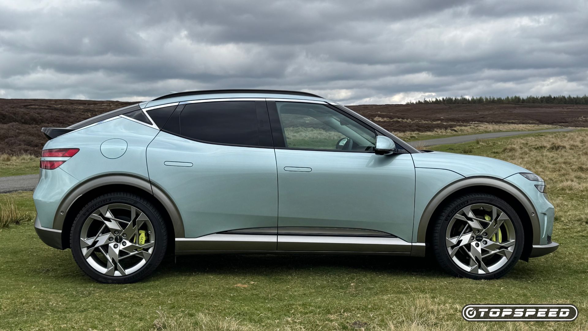 Static side profile shot of a mint green 2024 Genesis GV60 Sport Plus AWD parked on grass with hills and trees in the background.