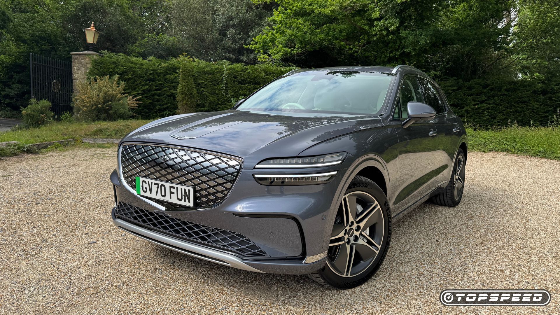 Static front 3/4 shot of a gray 2026 Genesis Electrified GV70 parked on gravel with trees in the background.