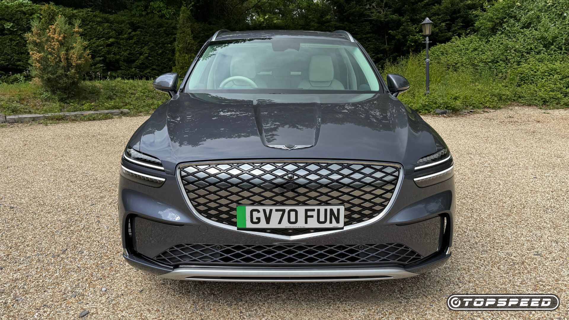 Static front-end shot of a gray 2026 Genesis Electrified GV70 parked on gravel with trees in the background.